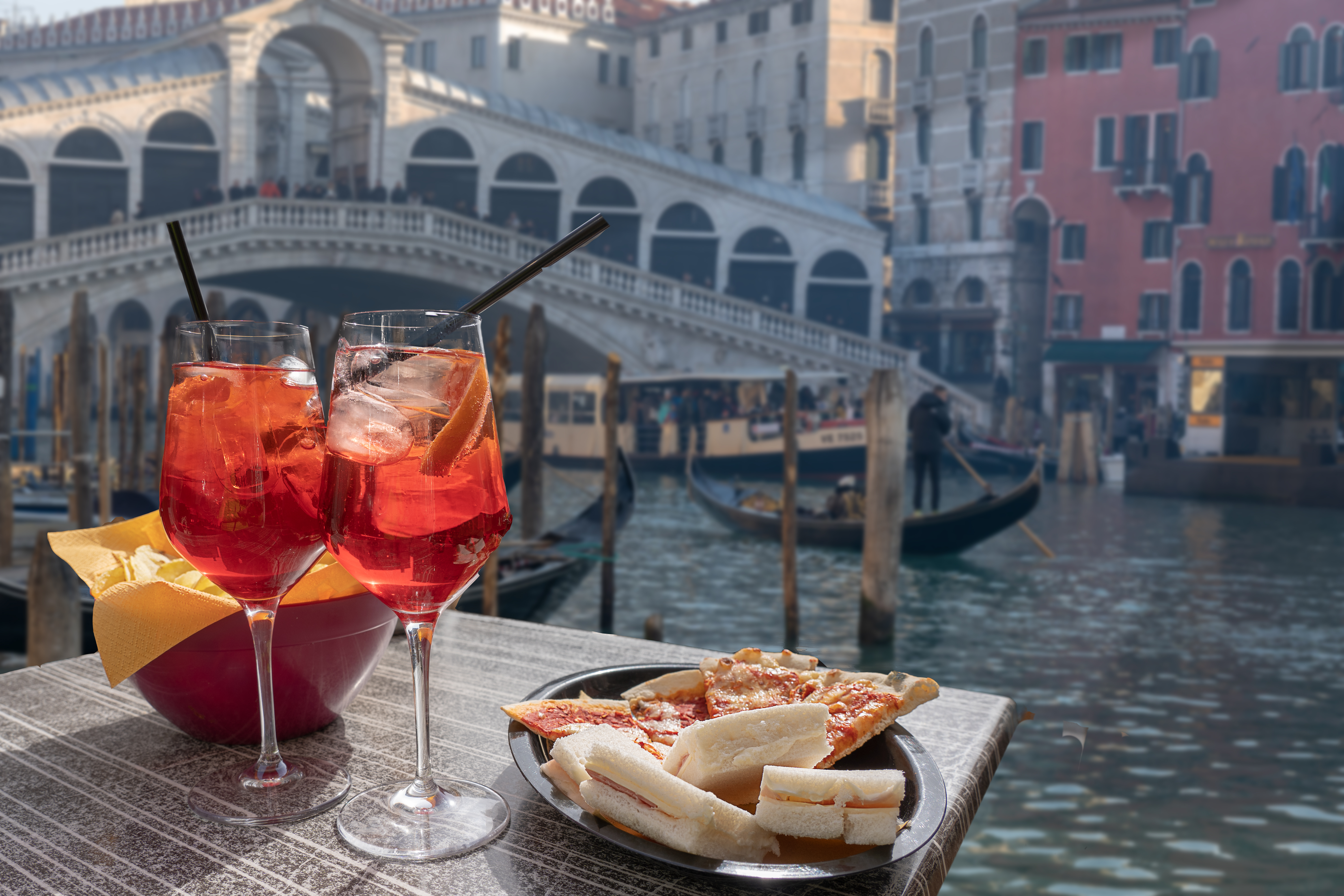 Cocktail for two prepared with straws and lemon and a bowl of snacks. In the background the Rialto Bridge in Venice.